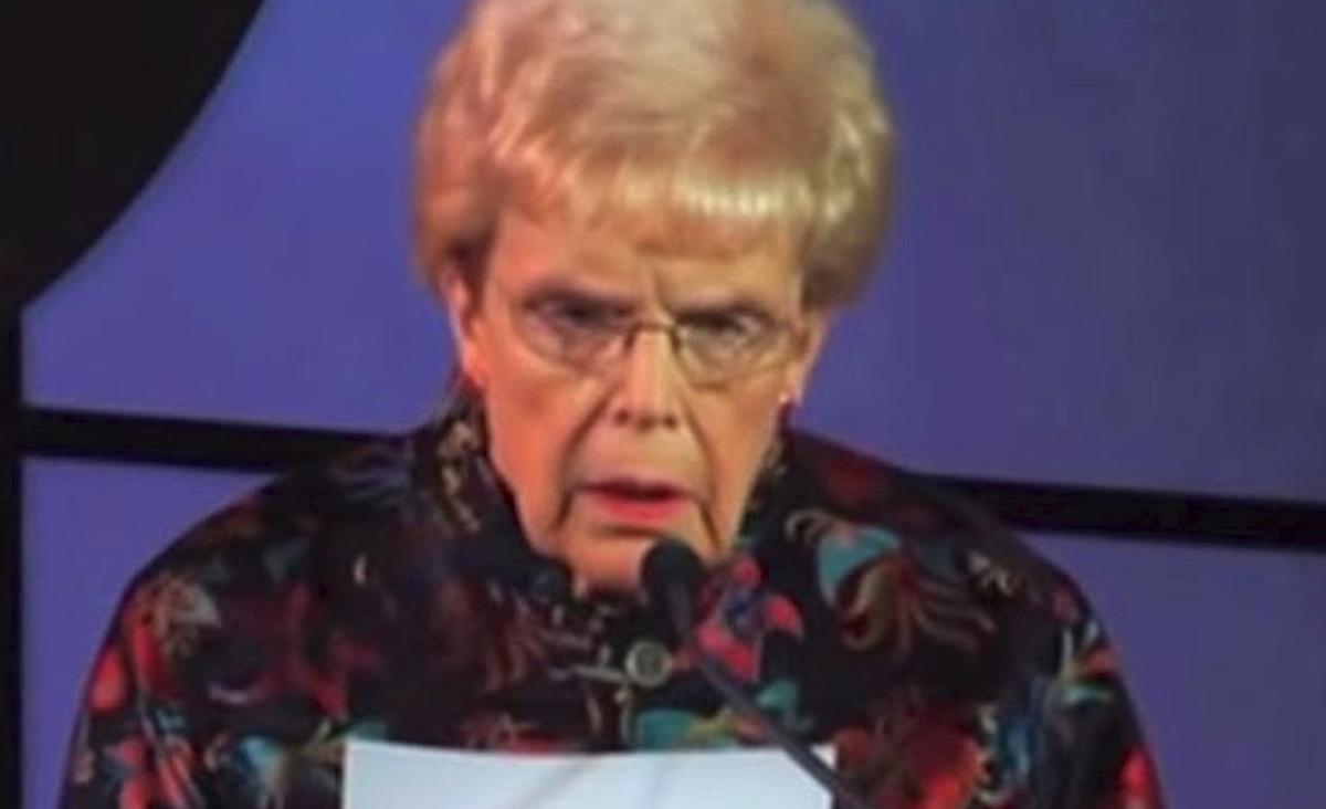 Mary Maxwell stands at a podium with a serious expression, wearing glasses and a colorful floral blouse, reading from a sheet of paper during a speech.