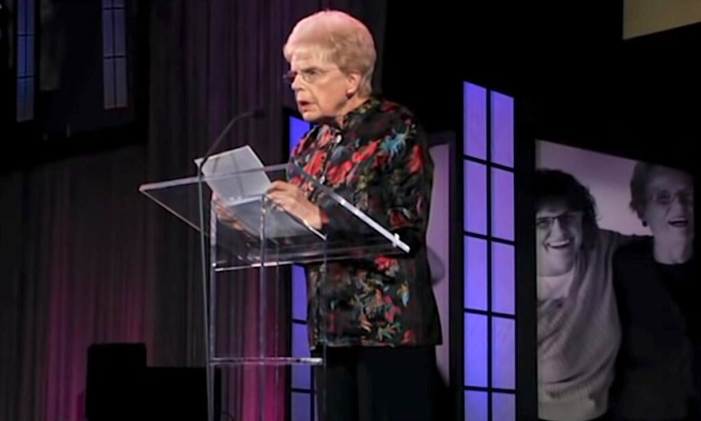 Mary Maxwell’s Hilarious Prayer About Getting Old 2 Mary Maxwell giving a speech at a transparent podium, wearing a colorful floral blouse and glasses, with a background image showing a smiling photo of herself.