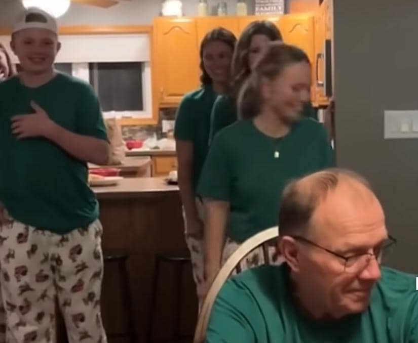 A group of four smiling young adults, all dressed in matching green shirts and patterned pajama pants, stand in a line in a cozy kitchen. In the foreground, an older man wearing glasses and a matching green shirt sits at a table, looking down with a gentle smile.