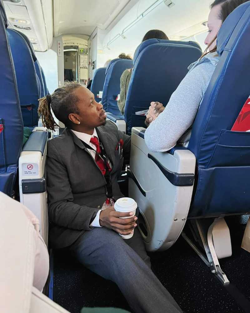 The image shows a male flight attendant sitting in an airplane aisle, holding a cup of coffee in one hand and gently holding a female passenger's hand with the other. The attendant is dressed in a uniform with a red tie and has dreadlocked hair. He is listening attentively to the passenger, who appears to be in conversation with him. The airplane seats are blue, and the overall atmosphere is calm and supportive.