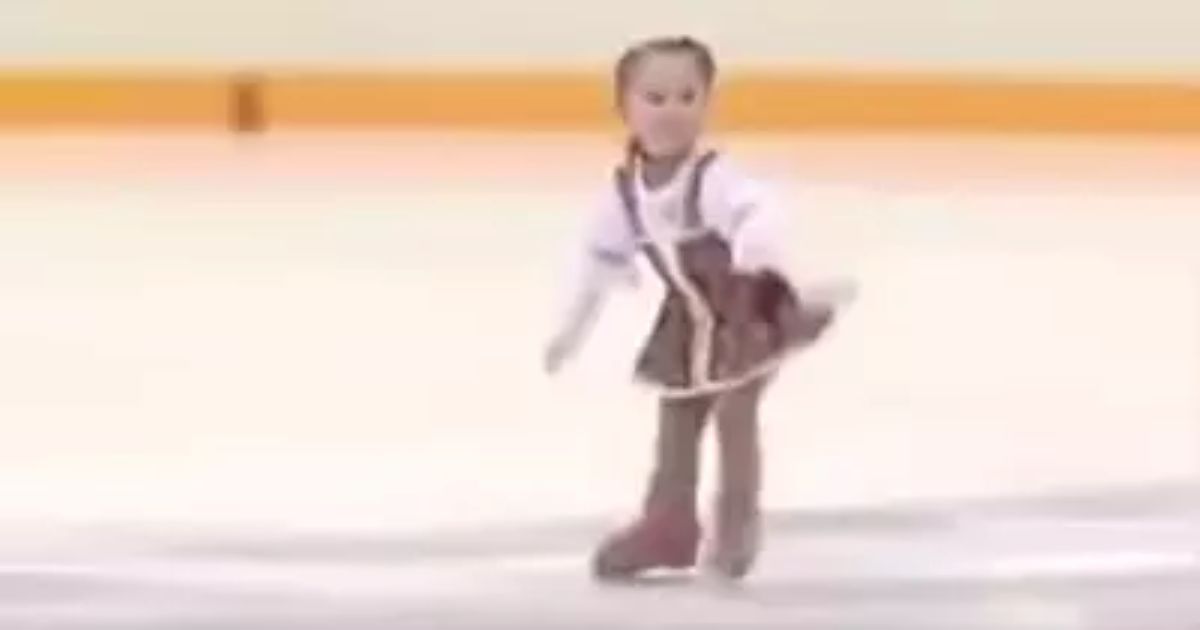 A young girl in a brown and white costume figure skating gracefully on an indoor ice rink.