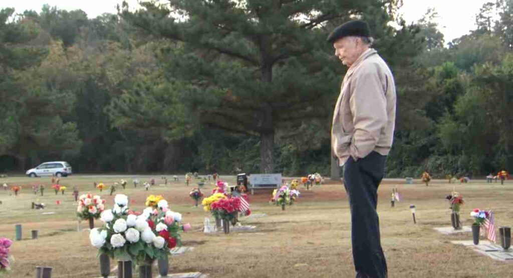 An elderly man stands solemnly in a cemetery, looking down at a grave adorned with colorful flowers. Tall pine trees and a white car in the background create a peaceful but somber atmosphere.