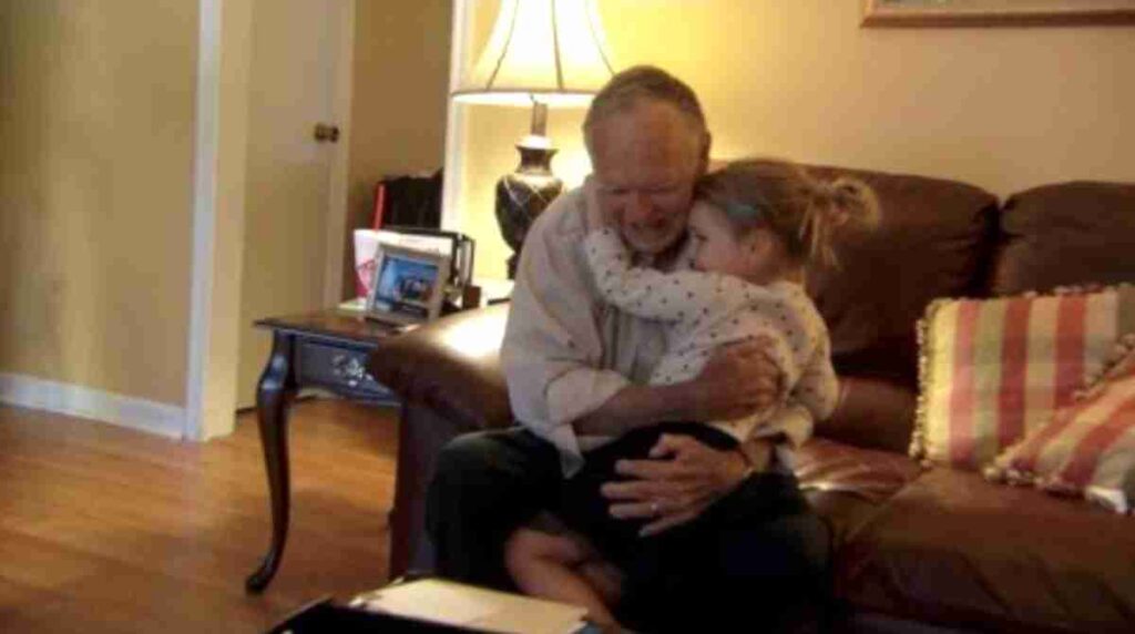 An elderly man sits on a couch warmly hugging a young girl who clings to him tightly. The cozy living room has soft lighting, a side table with family photos, and colorful pillows.