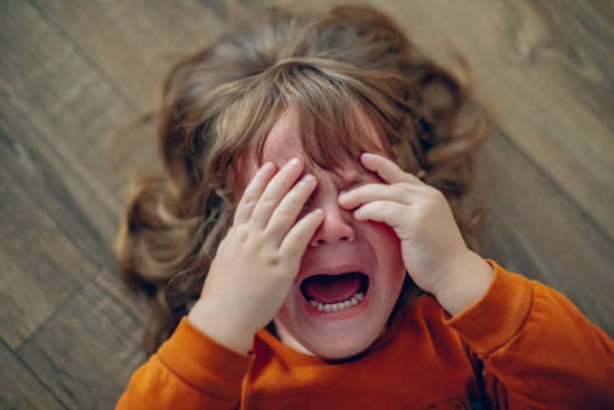 A young child with curly brown hair, wearing an orange sweatshirt, is crying with their mouth wide open and both hands covering their eyes while lying on a wooden floor.