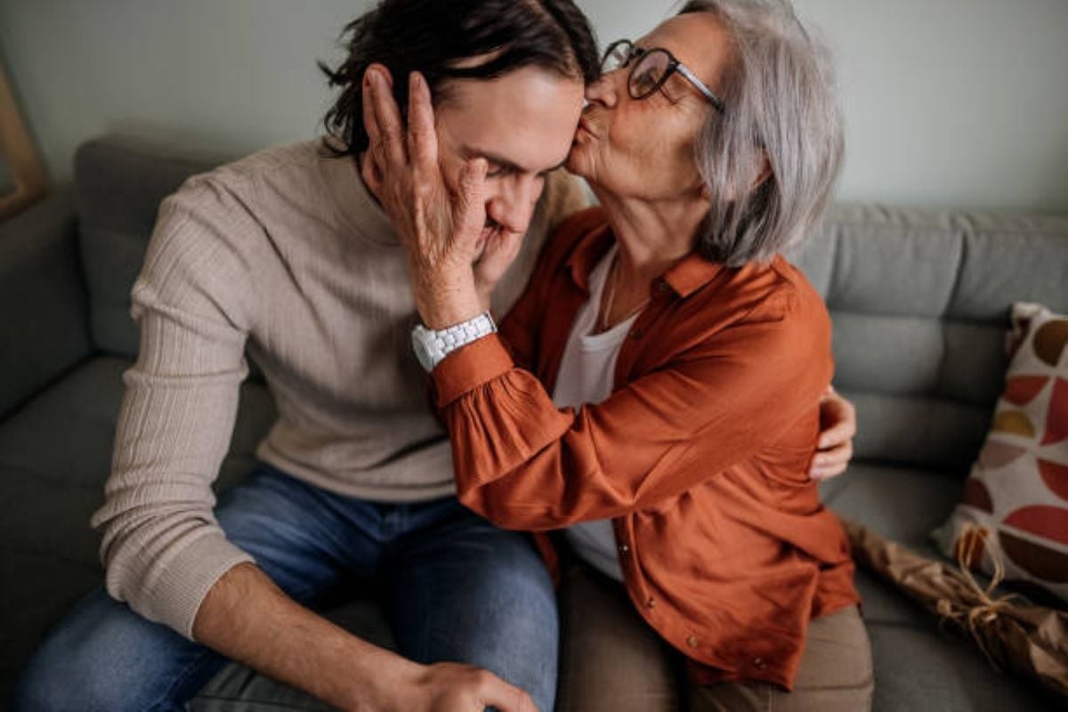 An elderly woman with gray hair and glasses gently holds a young man's face and kisses his forehead as they sit together on a couch, expressing love, comfort, and emotional support.