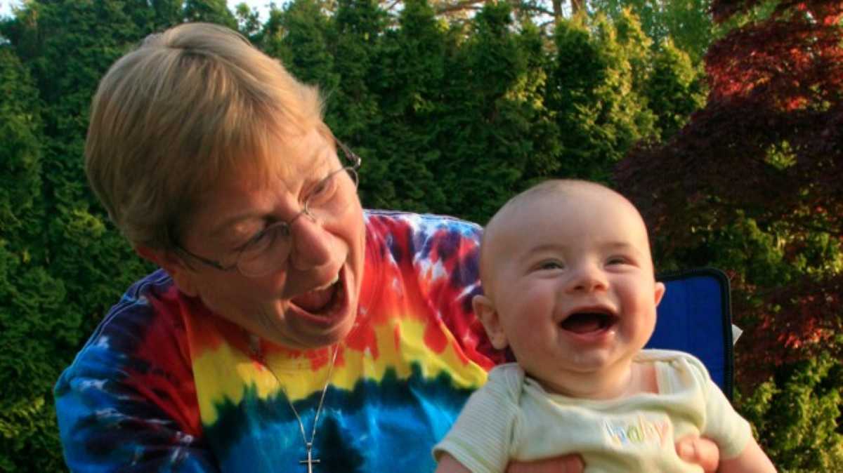 A joyful older woman in a tie-dye shirt laughing and holding a baby who is also laughing, in an outdoor setting with greenery in the background.
