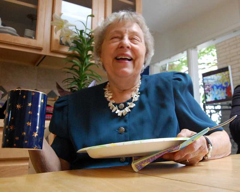 An elderly woman sitting at a kitchen table, laughing heartily with a coffee cup and a plate in front of her, in a well-lit room with a modern kitchen background.