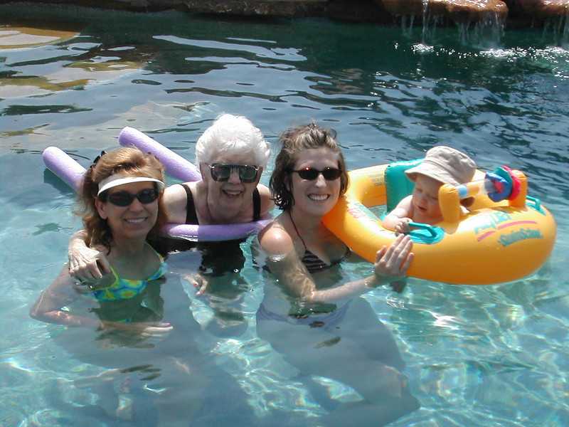 A family in a swimming pool including three adult women and a baby in a float, all smiling and enjoying the water.