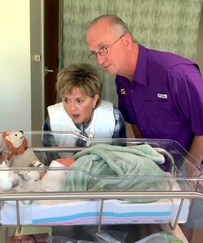 A grandmother and grandfather lean over a hospital bassinet, gazing in awe at a newborn baby. The grandmother looks surprised and emotional, while the grandfather appears touched and curious.