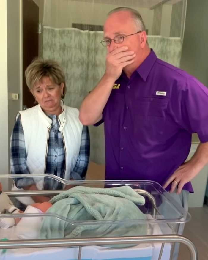 A grandmother and grandfather stand next to a hospital bassinet, looking emotional. The grandmother has a tender, touched expression while the grandfather covers his mouth in disbelief and awe.
