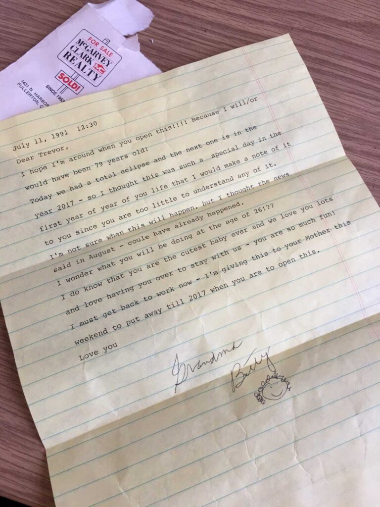 A full view of the same typed letter placed on a wooden desk, next to a torn envelope from McGarvey Clark Realty. The letter is signed "Grandma Betty" with a small smiley face drawing, and talks about a solar eclipse, Trevor's future, and the love she has for him.