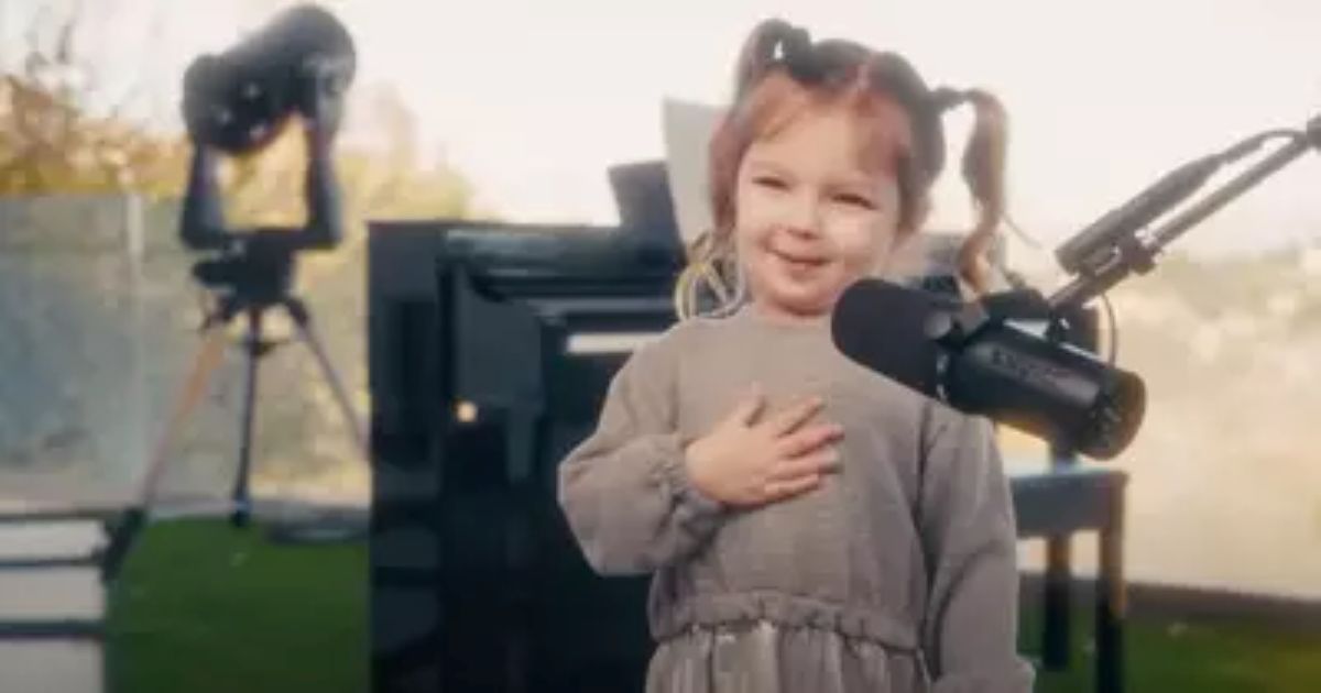 A young girl with light brown hair styled in pigtails stands in front of a microphone, smiling sweetly with one hand on her chest. She is wearing a gray dress and appears to be performing outdoors, with a piano and a telescope visible in the background. The scene is warmly lit, creating a heartwarming and peaceful atmosphere.