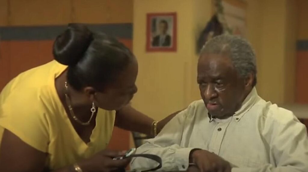 A woman in a yellow shirt leans toward an elderly man seated in a nursing home. The man looks downcast and unresponsive, with a blank expression on his face.