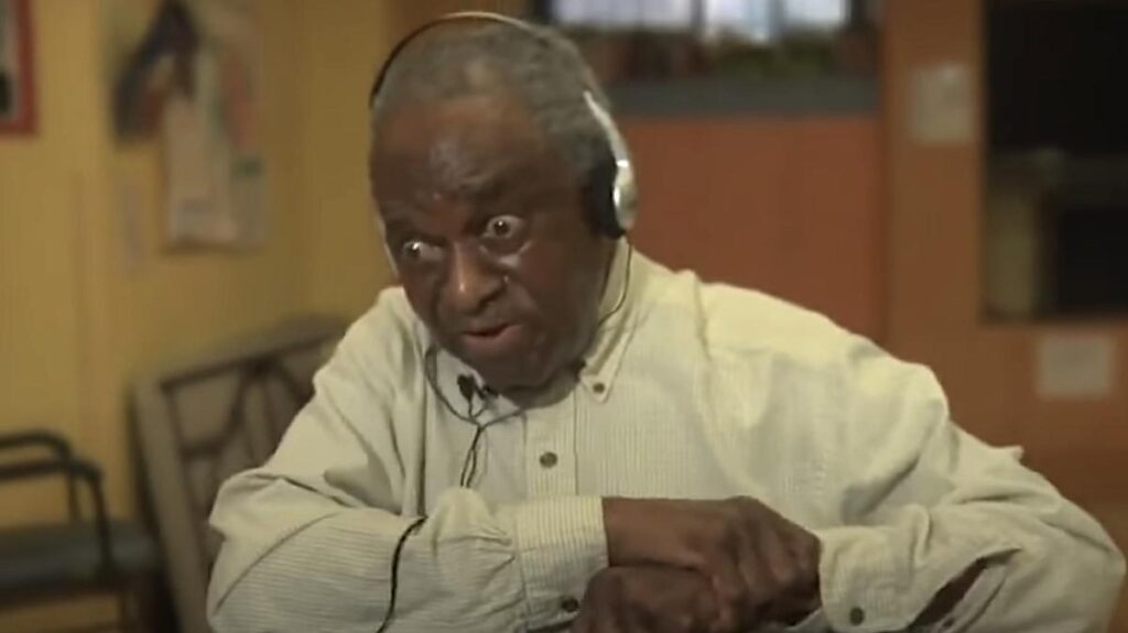An elderly man wearing headphones reacts with wide eyes and a lively expression as he listens to music in a nursing home. His body language shows surprise and engagement.
