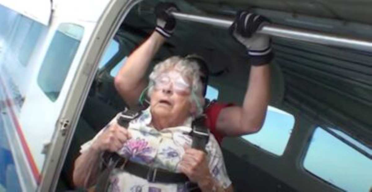 An elderly woman in a floral shirt, strapped in a tandem skydiving harness, stands at the open door of a plane with a skydiving instructor behind her, preparing for the jump.