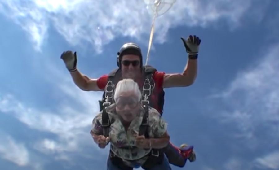 A senior woman skydives tandem-style with an instructor, both mid-air under a bright blue sky, with the wind rushing through her white hair and goggles on her face.
