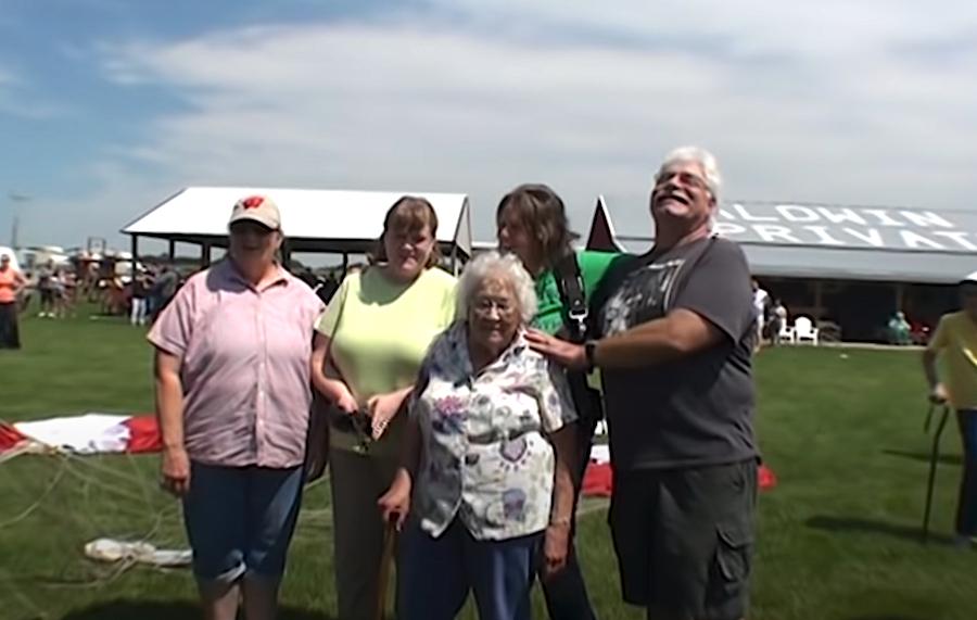 An elderly woman smiles while surrounded by supportive family members on the grass after completing her skydive, with the parachute and spectators visible in the background.