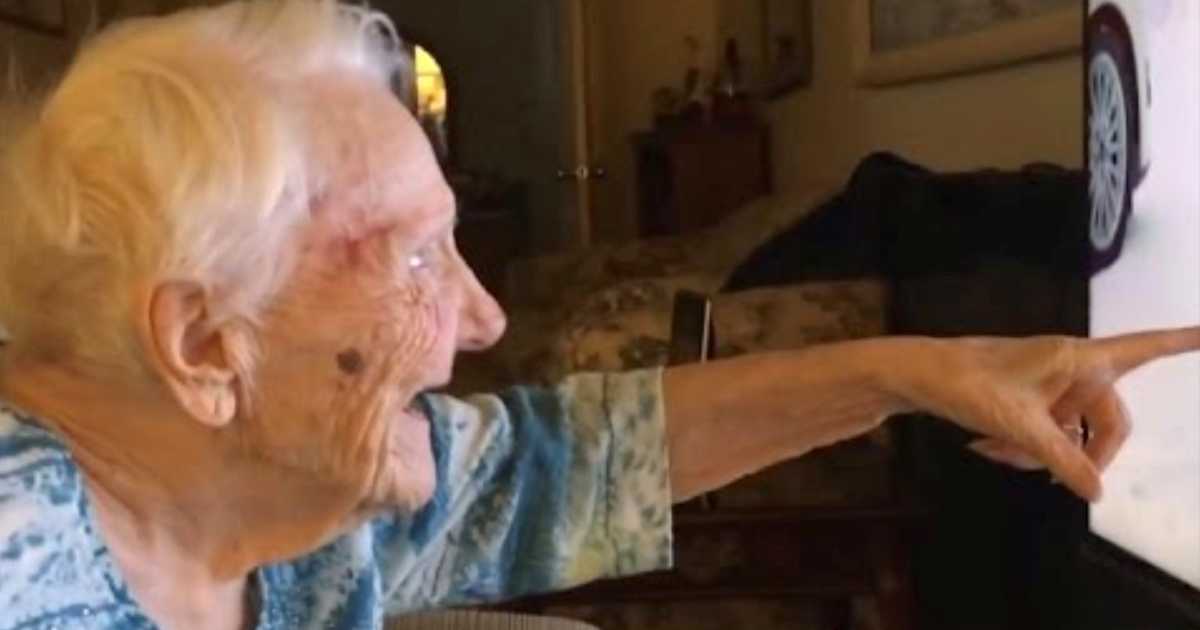 An elderly woman with white hair beams with joy as she points excitedly at a television screen. She sits in a cozy living room filled with warm lighting and a floral couch in the background.
