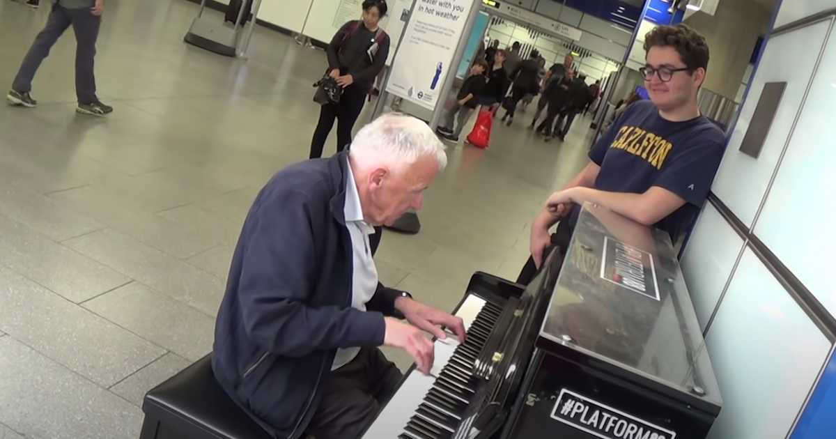 An elderly man plays a black upright piano in a busy airport terminal as a younger man in a navy t-shirt watches with a smile. Travelers pass by in the background, some stopping to observe.
