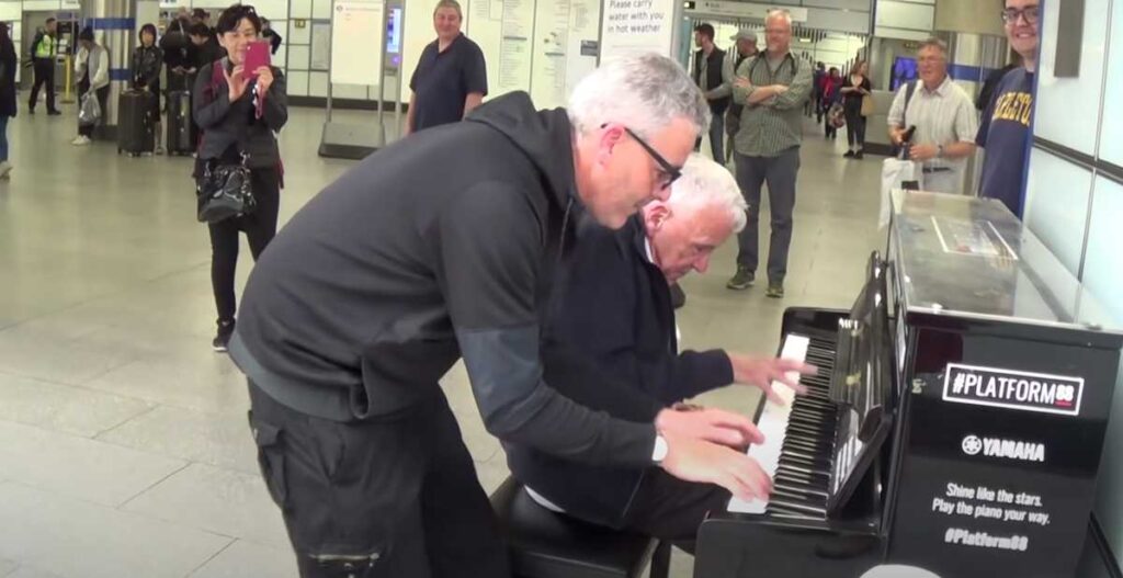 A younger man joins an elderly man at a black upright piano in a bustling airport. They play together while smiling onlookers gather, including a woman taking a photo with her phone.