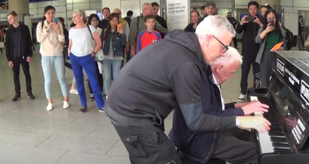 Two men, one elderly and one younger, share an upright piano in an airport terminal, playing in harmony. A growing crowd surrounds them, many recording the moment on their phones with smiles and awe.
