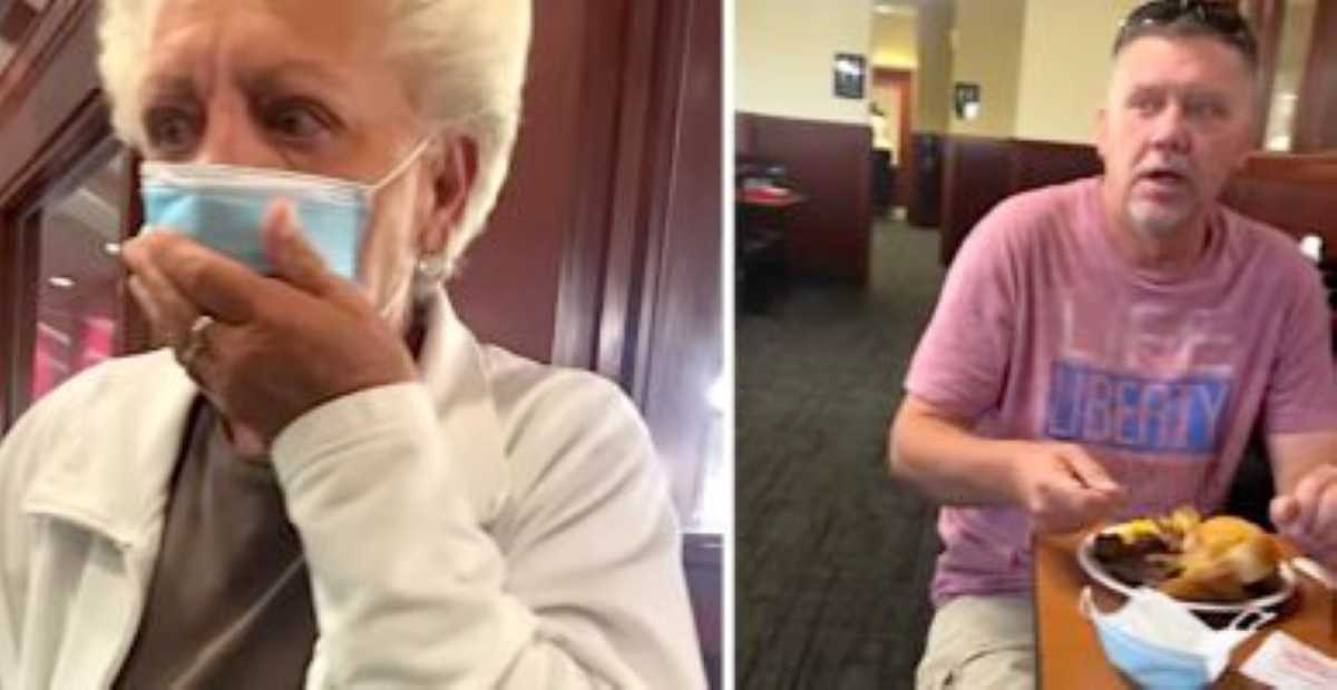 A grandfather wearing a face mask and white jacket covers her mouth in shock. Her eyes widen with emotion, suggesting a surprise moment in a restaurant.
