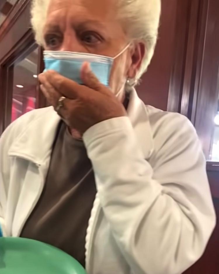 A grandfather wearing a face mask and white jacket covers her mouth in shock. Her eyes widen with emotion, suggesting a surprise moment in a restaurant. A grandfather wearing a “Liberty” t-shirt sits at a restaurant table mid-bite, staring in surprise. A mask lies on the table, and his plate is full of food.