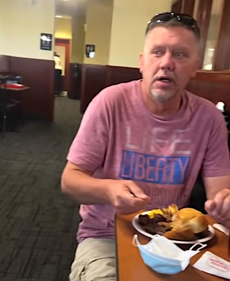 A grandfather wearing a “Liberty” t-shirt sits at a restaurant table mid-bite, staring in surprise. A mask lies on the table, and his plate is full of food.