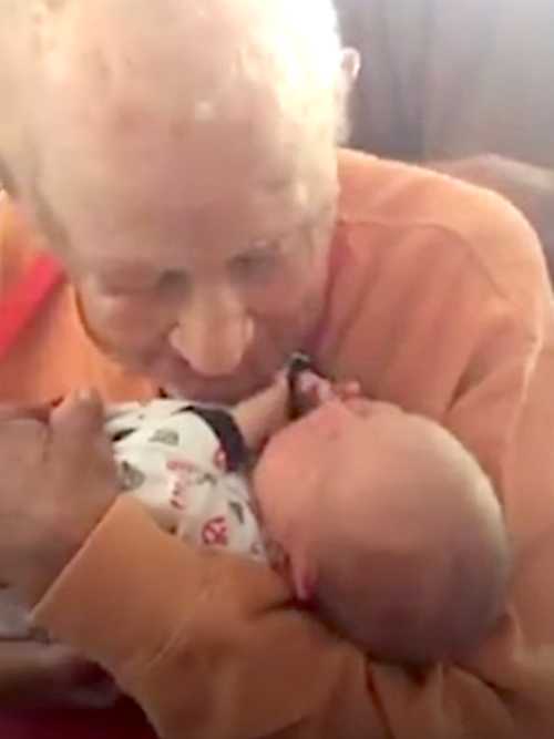 A touching close-up of a 105-year-old man smiling gently as he cradles a newborn baby in his arms. The baby looks up at him, and the two share a quiet, heartwarming connection.