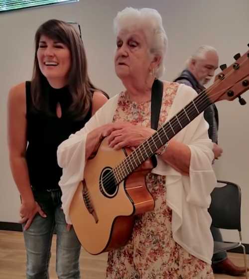An elderly woman in a floral dress and white cardigan holds a guitar while standing next to a smiling younger woman. They are inside a room with chairs in the background.