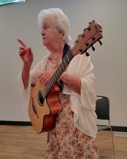 An elderly woman wearing a floral dress and white cardigan holds a guitar and raises her finger while speaking, as if making a point or telling a story.