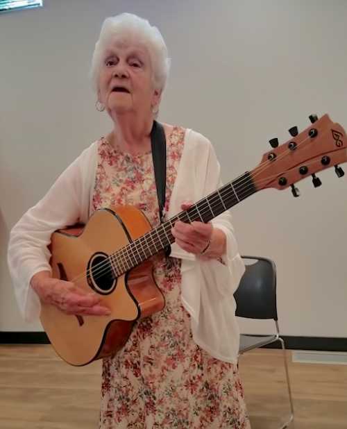 An elderly woman in a floral dress and white cardigan sings and plays a guitar, looking expressive and full of joy during her performance.