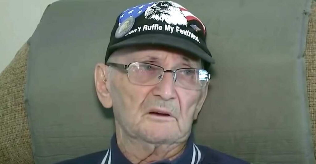 A close-up of the elderly man sitting on a couch, looking solemn and reflective while wearing glasses and the same patriotic cap.