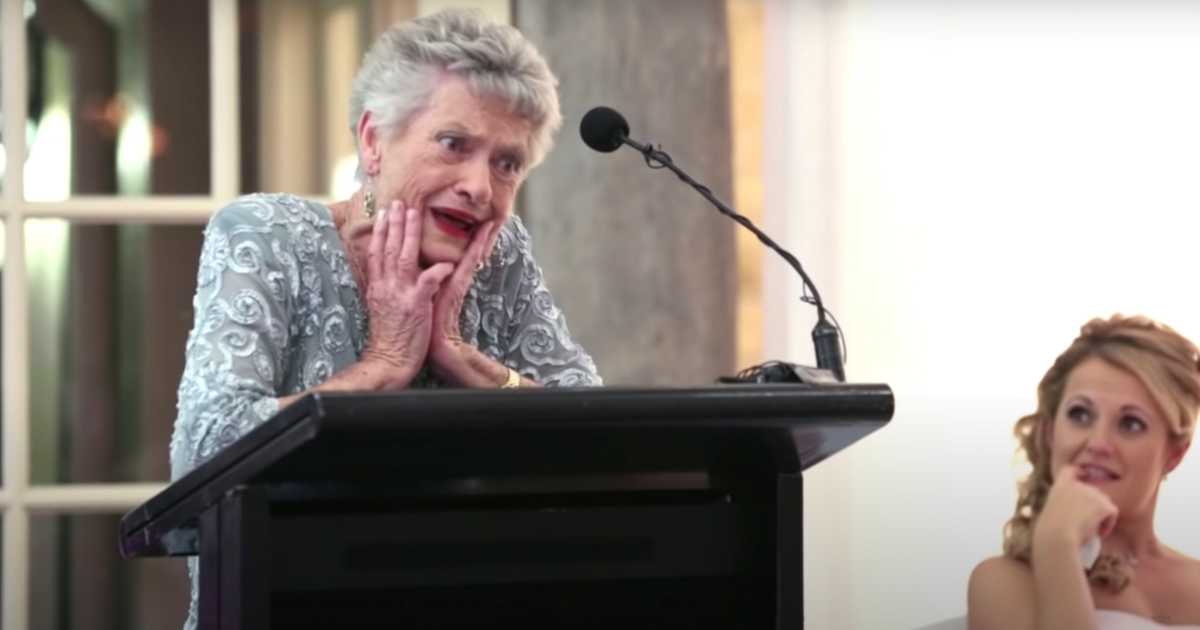 An elderly woman in a light blue embroidered dress stands at a podium with a microphone, holding her face with both hands in a playful, dramatic expression. A smiling blonde woman in a wedding dress sits nearby, watching her with amusement. The setting appears to be a wedding reception.