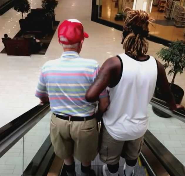 A young man with blonde-tipped dreadlocks and a sleeveless shirt stands beside an elderly man wearing a red cap and striped polo, holding his arm as they ride down an escalator together in a shopping mall.