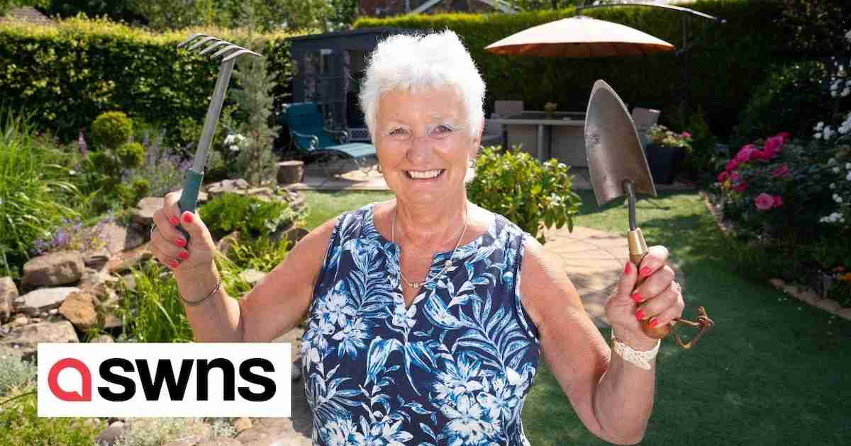 A smiling elderly woman with short white hair holds gardening tools in both hands while standing in a beautifully landscaped backyard. She wears a sleeveless floral top and has bright red nail polish. Behind her, the garden features lush greenery, flowers, and a patio area with outdoor furniture.
