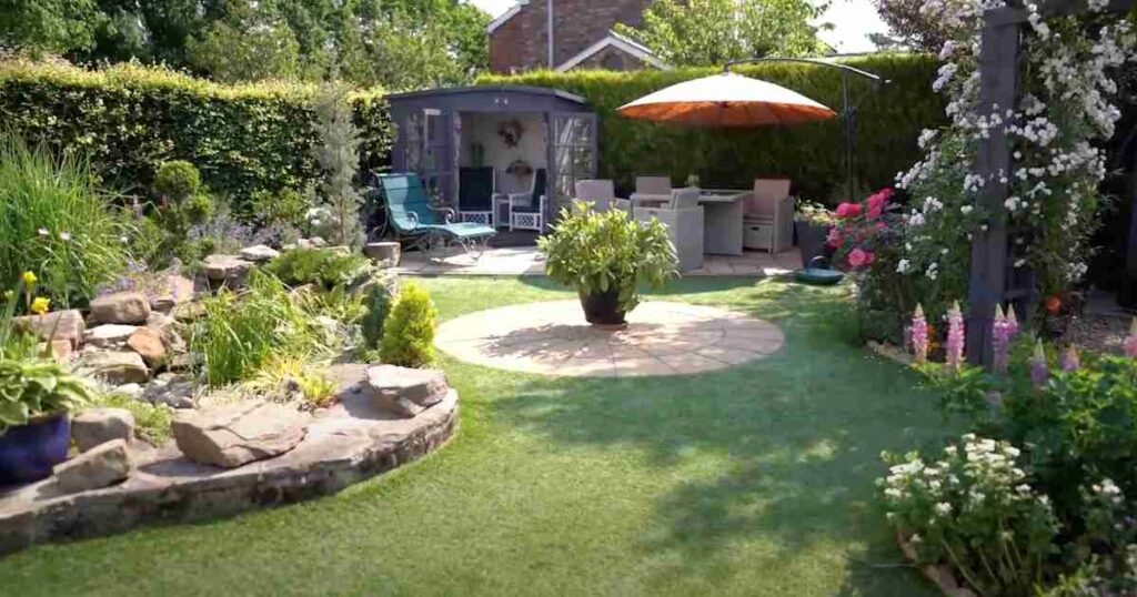 A vibrant backyard garden with neatly trimmed grass, a stone-lined flower bed, blooming flowers, and a variety of plants. A circular patio area with a large potted plant sits in the center, while a cozy outdoor seating area with a parasol and garden furniture is visible in the background.