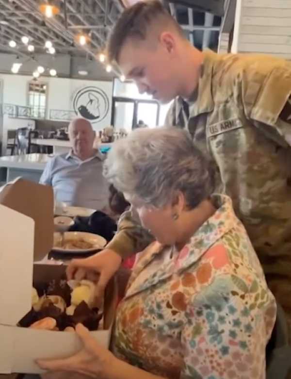 A close-up of a joyful surprise at a restaurant—an elderly woman examines a box of cupcakes while her grandson, dressed in a U.S. Army uniform, stands behind her, moments before she realizes he's there.