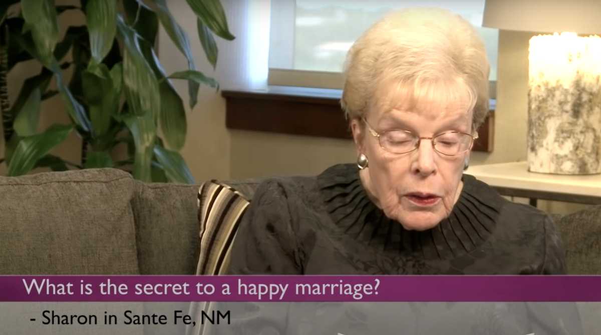 An elderly woman with short blonde hair and glasses, wearing a black blouse, reads aloud while sitting on a couch. A question appears on screen: “What is the secret to a happy marriage?” – Sharon in Santa Fe, NM. A houseplant and a lit candle are visible in the background.
