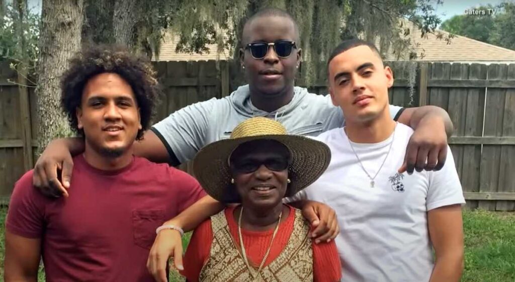 A cheerful outdoor family photo showing three young men standing behind their elderly grandmother. She wears a sunhat, sunglasses, and a red top, while the men smile with their arms affectionately around her in a backyard setting.