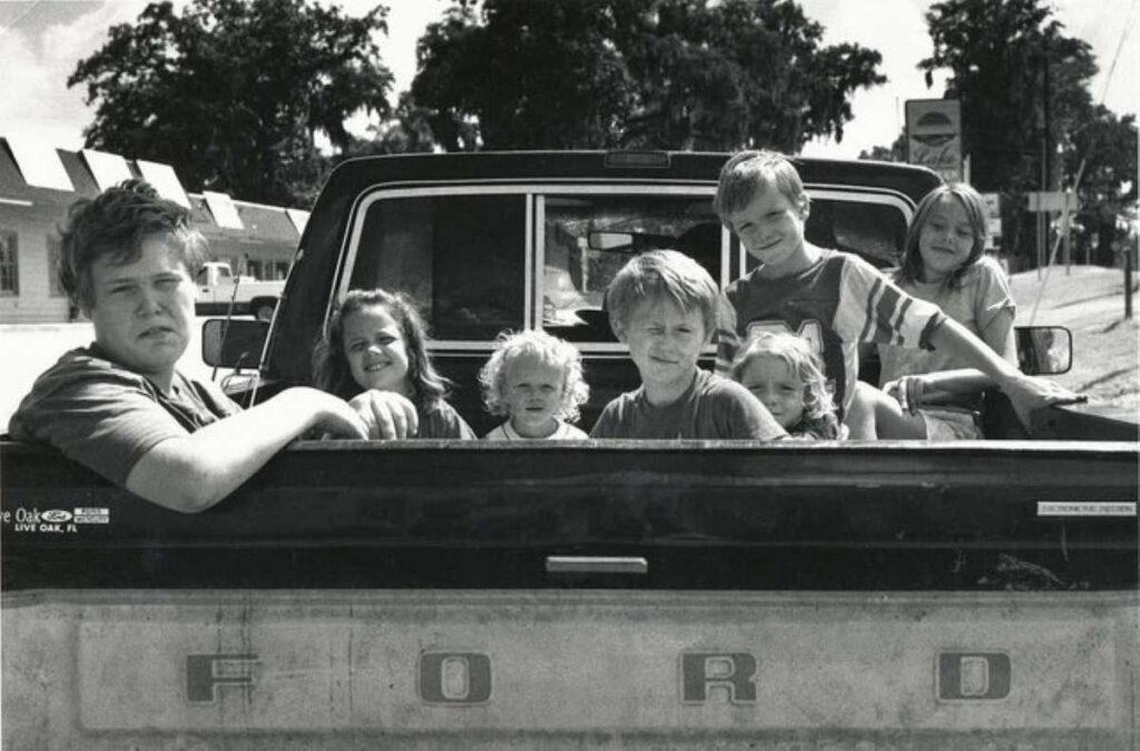 Seven children sitting in the back of a Ford pickup truck on a sunny day, captured in a vintage black-and-white photo.