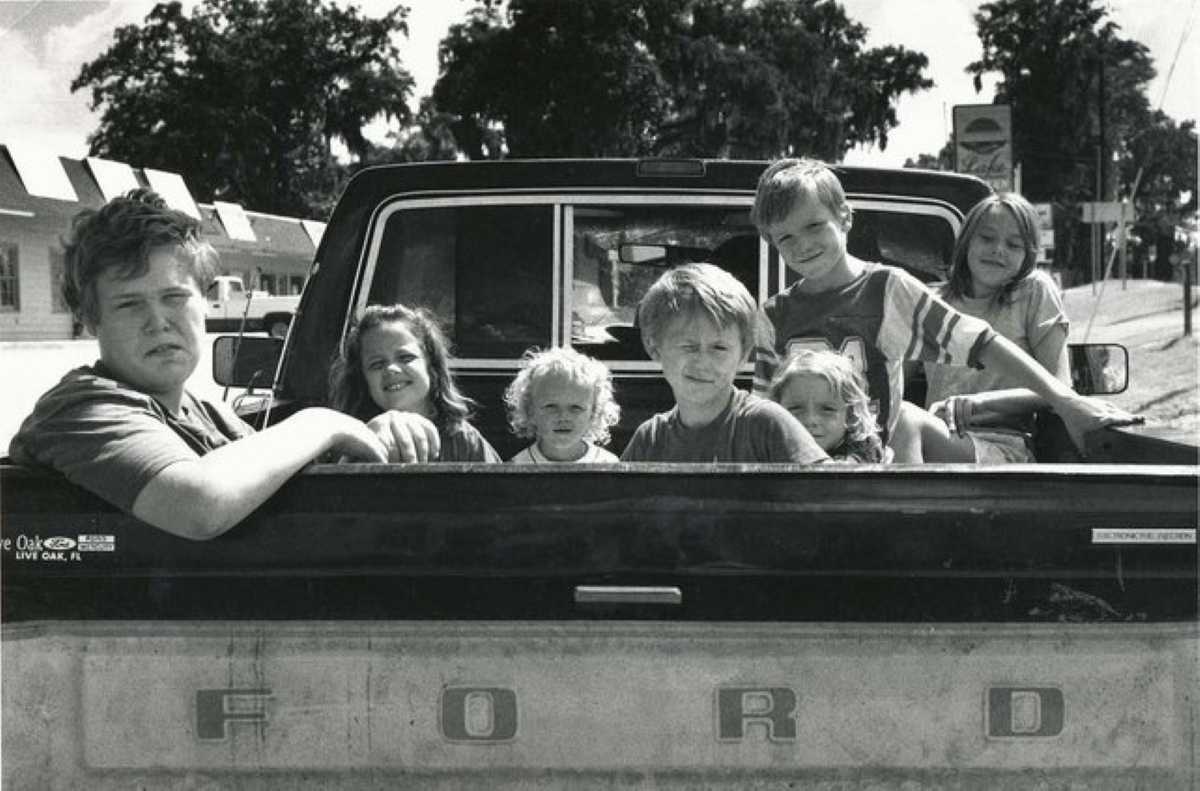 Seven children sitting in the back of a Ford pickup truck on a sunny day, captured in a vintage black-and-white photo.