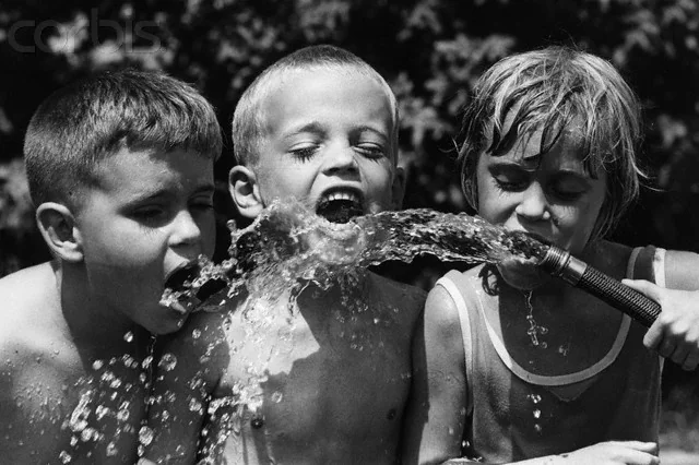 Three young kids drinking from a garden hose, laughing and enjoying a hot day in a classic black-and-white summer scene.
