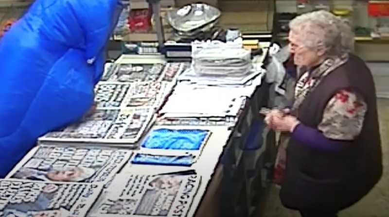 Surveillance footage of an elderly woman standing calmly across the counter from a person in a large blue hooded coat, believed to be a robber, inside a convenience store filled with newspapers and magazines.