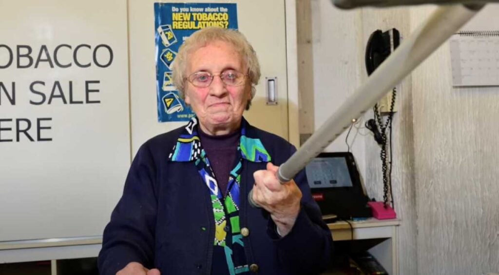 Close-up of an elderly woman smiling slightly and holding her cane out toward the camera in a mock-defensive pose, standing in front of a tobacco sales sign and a cash register inside her shop.