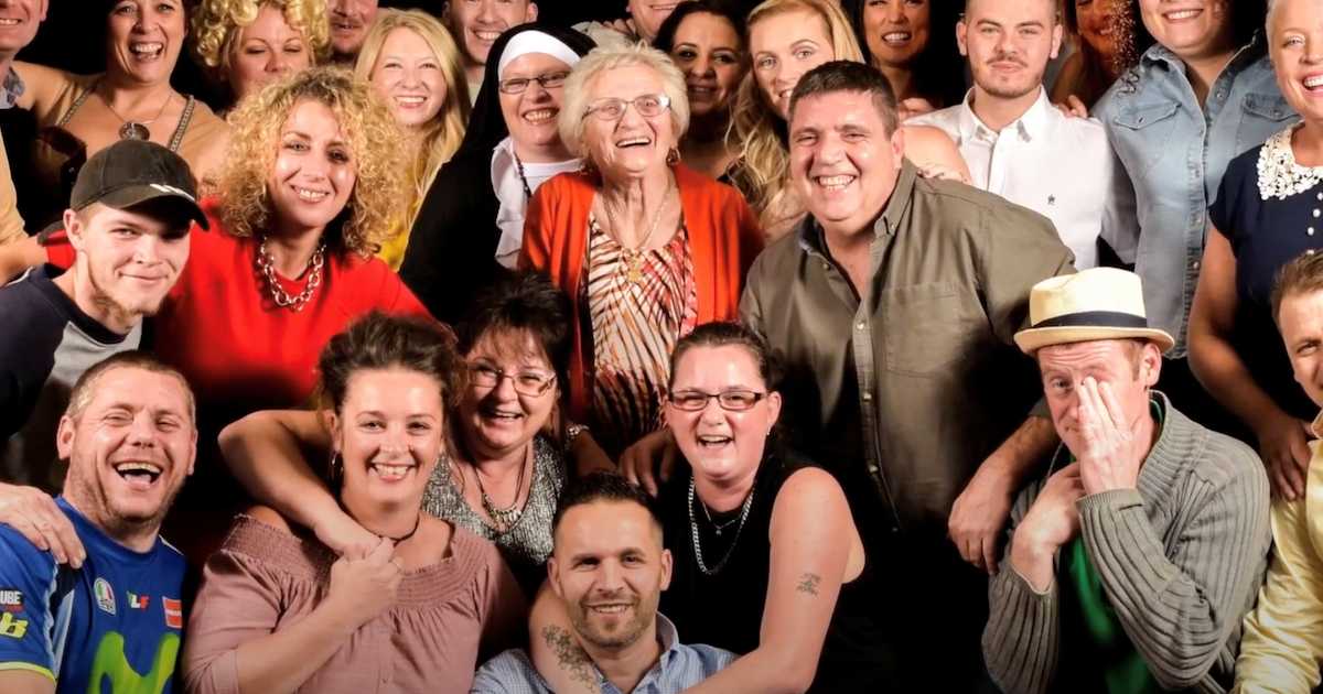 A joyful multigenerational family group photo featuring dozens of smiling people gathered around an elderly woman in the center, wearing a red cardigan. The crowd includes people of various ages, laughing and embracing, showing a strong sense of love and connection.