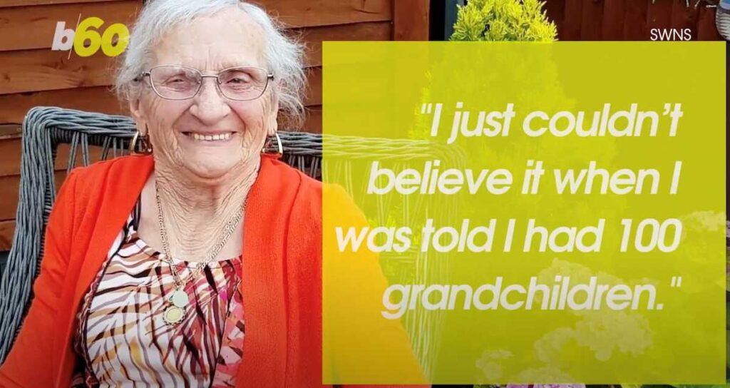 A smiling elderly woman with white hair and glasses sits outside on a wicker chair, wearing a patterned dress with a bright orange cardigan and gold jewelry. A yellow text overlay quotes her saying: “I just couldn’t believe it when I was told I had 100 grandchildren.” The image includes the “b60” and “SWNS” logos.