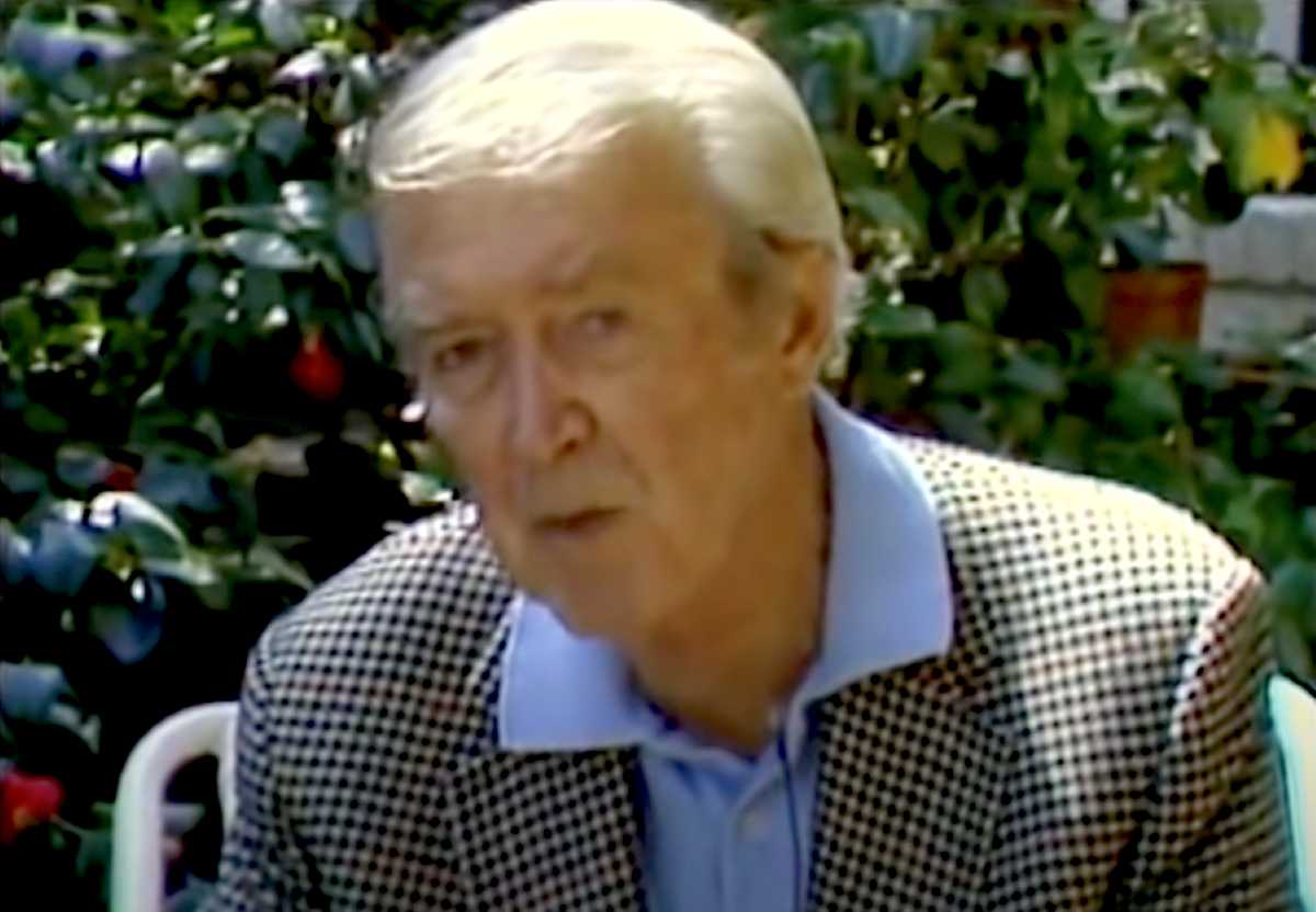 An elderly Jimmy Stewart sits outdoors in front of lush greenery, wearing a checkered blazer over a light blue collared shirt, speaking directly to the camera.