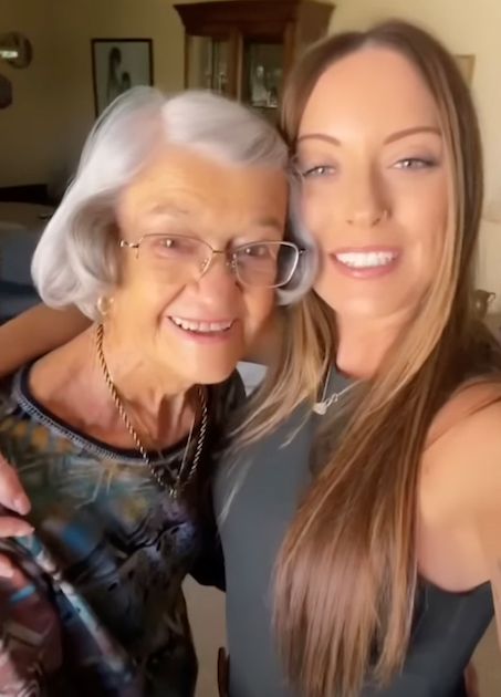 A joyful elderly woman and her granddaughter pose for a close-up selfie indoors, both smiling warmly at the camera.