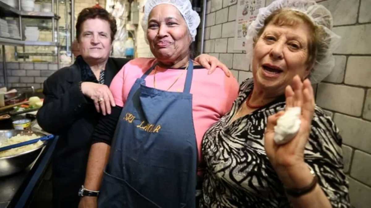 Three older women smile warmly in a cozy kitchen at Enoteca Maria, a New York restaurant run by women over 50 from around the world. One wears an apron labeled "Sri Lanka," while the others stand beside her in hairnets and casual attire, exuding pride and joy. The background shows a tiled wall, shelves with dishes, and food preparation in progress.